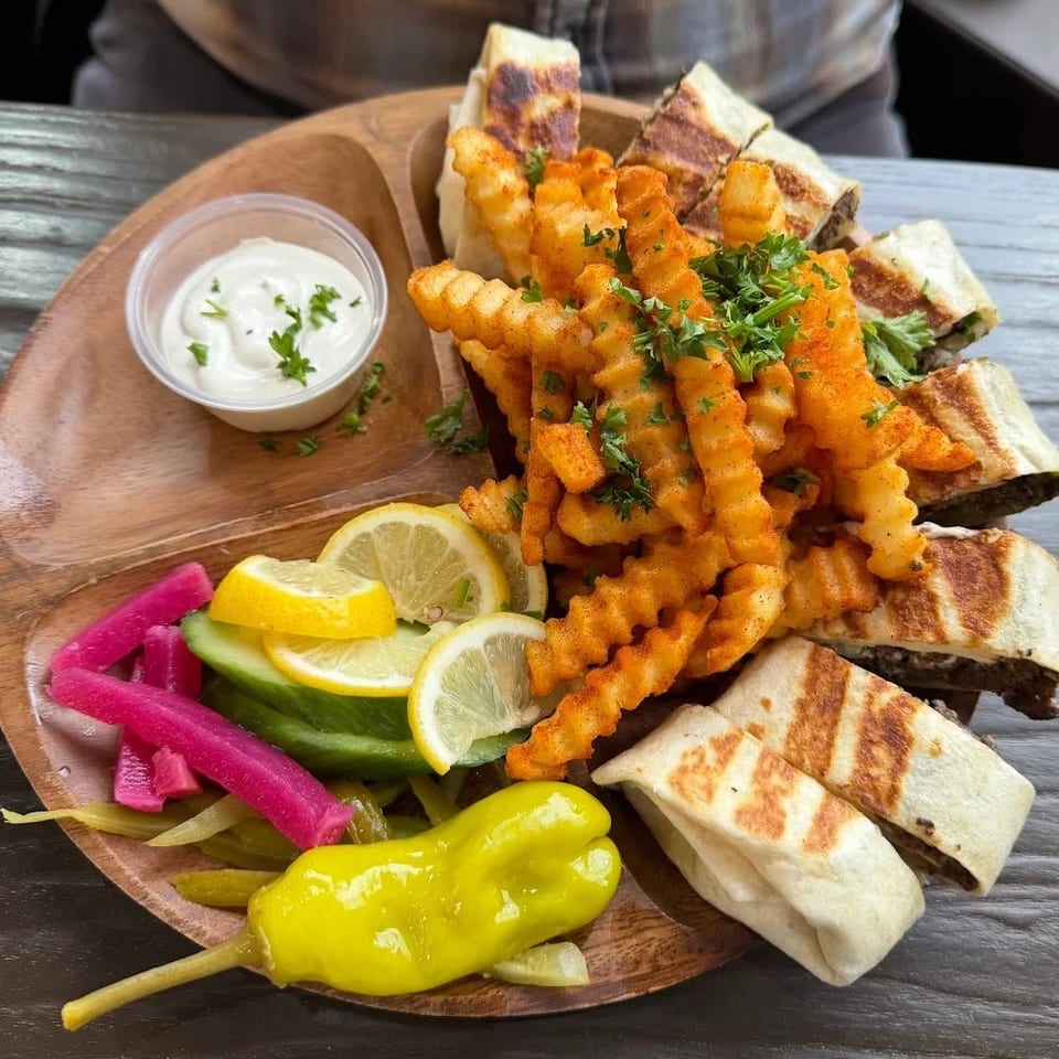 A wooden platter with shawarma wraps, crinkle-cut fries, pickles, lemon slices, and a side of sauce.