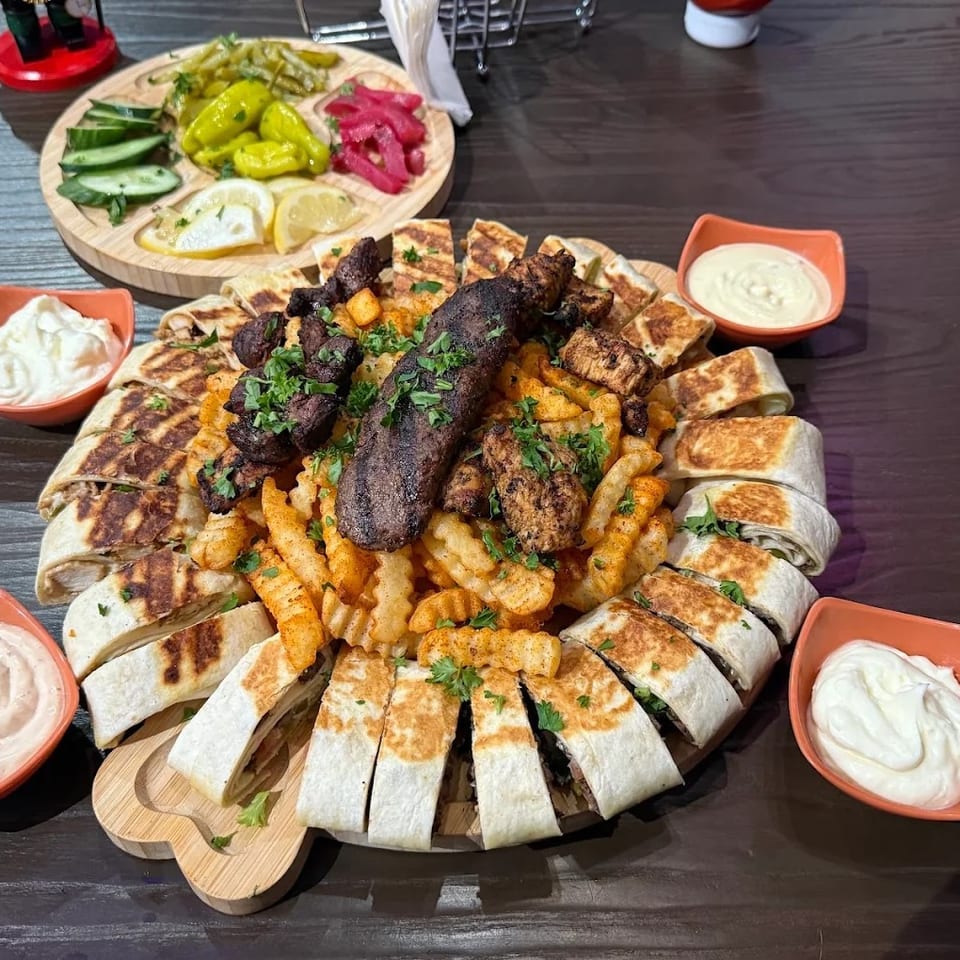A large platter of shawarma wraps, fries, grilled meats, and dipping sauces on a wooden table.