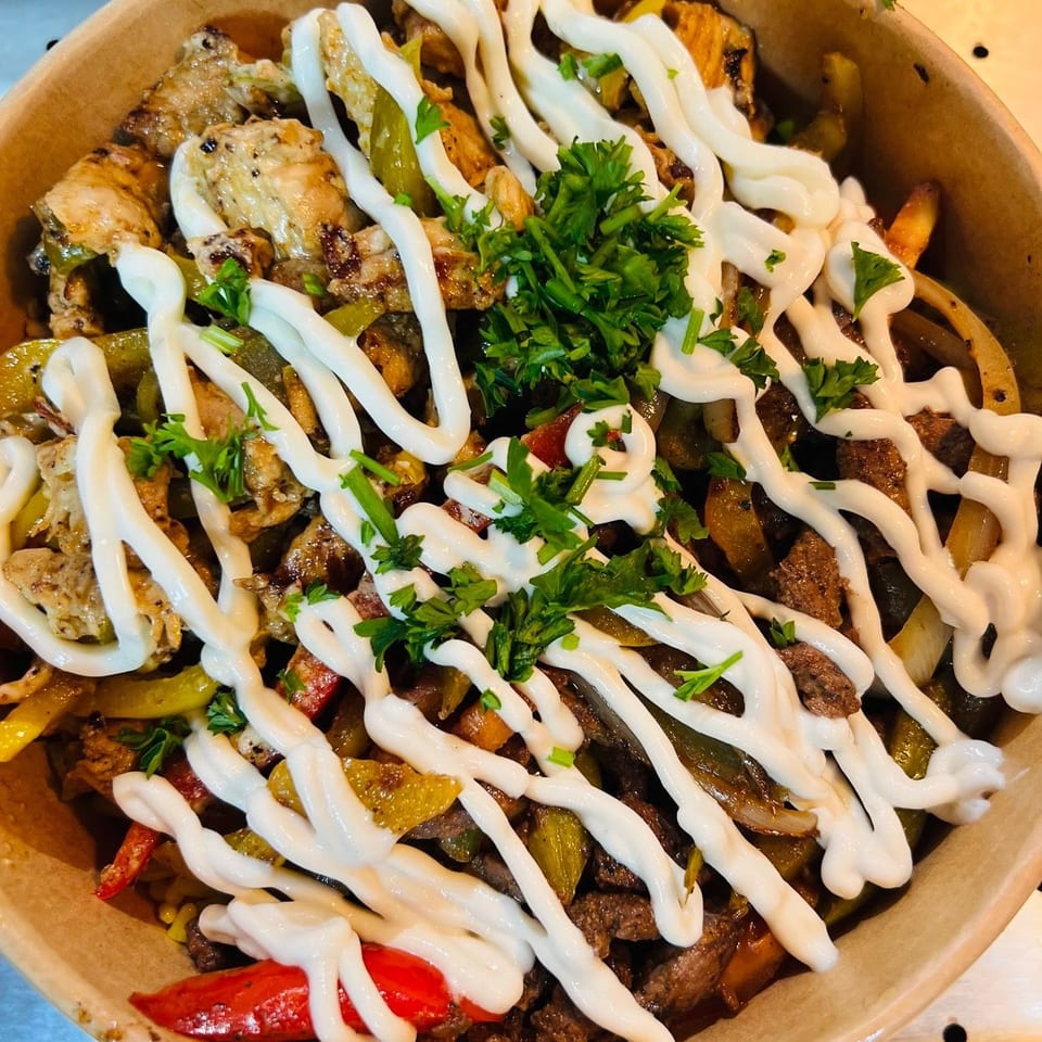 Close-up of a bowl filled with grilled chicken and beef, peppers, onions, drizzled with white sauce and parsley.